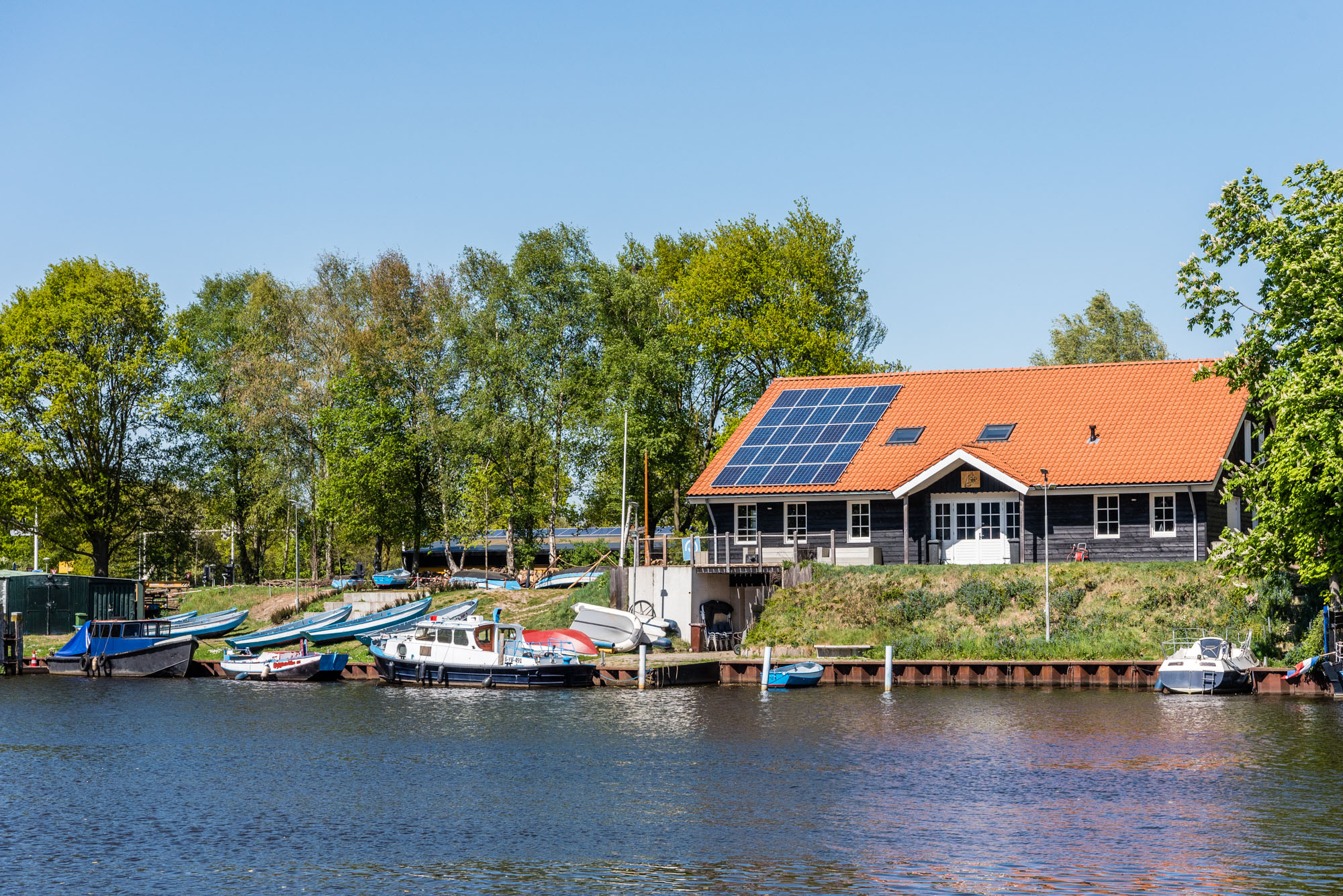 gebouw van de Karel Doormangroep vanaf de overkant van het water gezien met boten op de wal, veel groen rondom en zonnepanelen op het dak